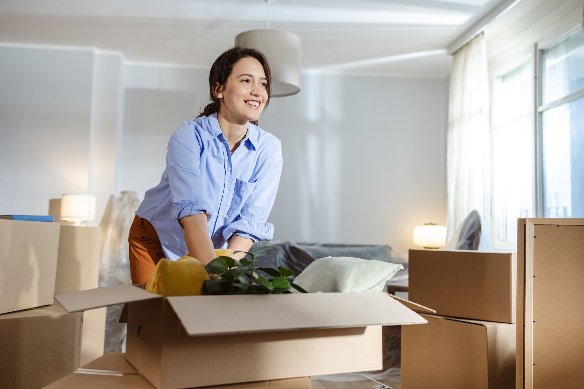 Smiling positive young woman surrounded by boxes while moving, unpacking personal belongings